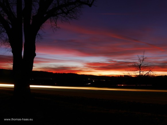Bild des Monats, März 2024 - ein Sonnenuntergang als Langzeitbelichtung mit Fahrzeuglichtern im Vordergrund.